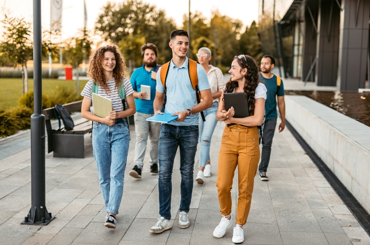 A group of smiling students walking outdoors on a college campus, carrying books and backpacks, enjoying a sunny day.