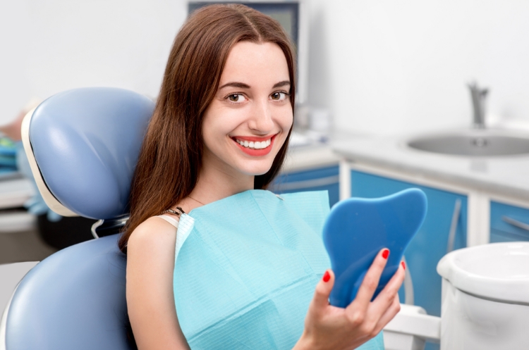 A smiling woman sitting in a dental chair, holding a blue dental mirror. She is wearing a dental bib and appears satisfied with her dental care in a clean, modern dental office.